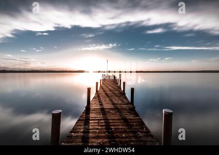 Un molo di legno sul lago Starnberg in Baviera getta lunghe ombre nel luminoso tramonto sotto un cielo leggermente nuvoloso. Il cielo si riflette nel lago liscio. Foto Stock