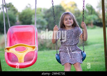 hermosa niña rubia de 8 años disfruta de la naturaleza en un escenario al aire libre lleno de verdor y luz mientras se columpia con gracia Foto Stock