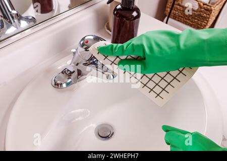 Una persona con guanti protettivi in gomma verde che pulisce un rubinetto in bagno, vista ravvicinata Foto Stock
