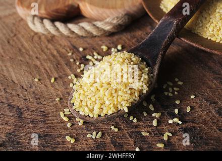 Asciugare Bulgur di grano in cucchiaio di legno rustico sfondo. Eventuali fuoriuscite di bulgur Foto Stock