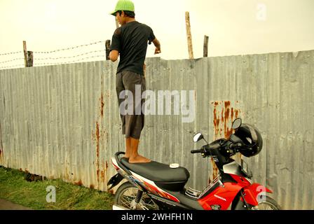 Un pescatore ricreativo guarda dietro mentre si trova su una moto parcheggiata, mentre pesca da uno stagno situato dietro un confine recintato di un cantiere navale nel villaggio costiero di Marunda, Cilincing, Giacarta settentrionale, Indonesia. Foto Stock