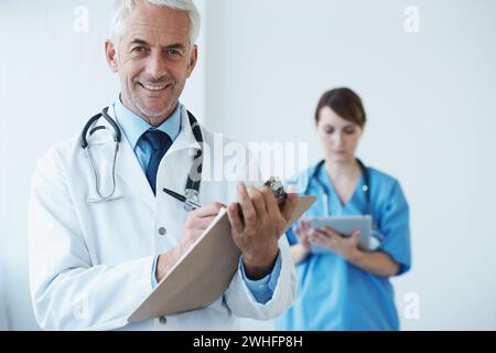 Ritratto, medico maturo che scrive sugli appunti e con un infermiere che naviga su un tablet digitale sullo sfondo. Medico e infermiere senior in piedi Foto Stock