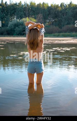 La vista posteriore di una donna in piedi nel fiume si toglie la camicia Foto Stock