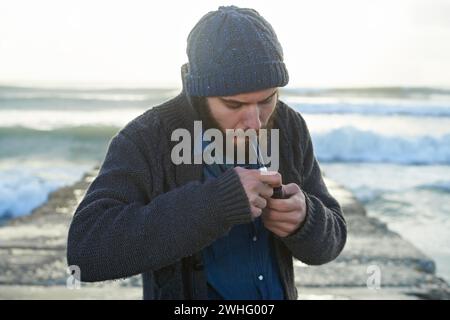 Barbuto, uomo e fumare una pipa vicino all'oceano, abitudine più leggera e tabacco la mattina d'inverno per l'alba. Ragazzo inglese, nicotina e fumatore vintage per la calma Foto Stock