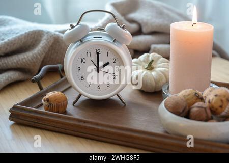 Sveglia vintage bianca, candela, decorazioni autunnali e dolci su un comodino con coperta naturale di lana, concetto di ripiegamento Foto Stock