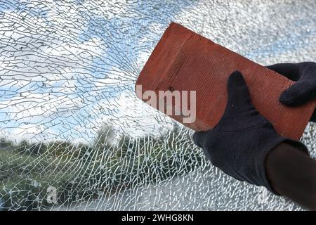 Le mani con i guanti e i mattoni distruggono una finestra in vetro di sicurezza laminata, il concetto di violenza e vandalismo, lo spazio di copia Foto Stock