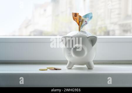 Salvadanaio con banconote e monete in euro su davanzale, concetto di thrift durante l'inflazione, spazio di copia, focus selezionato Foto Stock