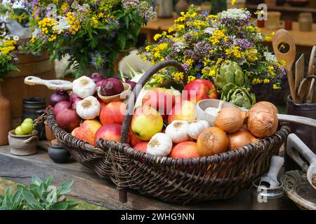 Cesto pieno di mele, cipolle, aglio e carciofi, raccolto dall'orto decorato con mazzi di fiori selvatici Foto Stock