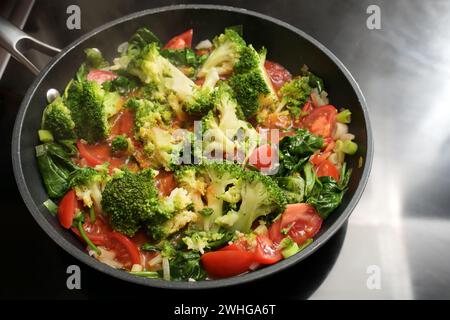 Padella di verdure con broccoli, pomodori, cipollotti e spinaci sul piano cottura, cucinando cibi vegetariani sani con ingrossati freschi Foto Stock