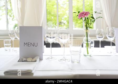 Elegante ambiente da tavola, posto con menu a carte, bicchieri, posate, tovagliolo e un fiore rosa su una tovaglia bianca per una cena festosa Foto Stock