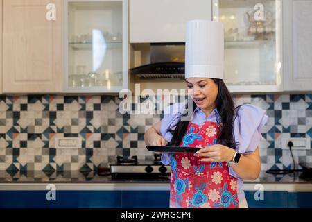 Bella donna indiana vestita in uniforme da cuoca con grembiule, cappello da cuoco in piedi in cucina con teglia antiaderente e si eccita dopo aver guardato h Foto Stock