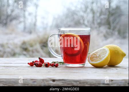 Tè caldo da rosa rossa fianchi con fette di limone in una tazza di vetro all'aperto su un rustico tavolo di legno, bevanda calda sana e calda aga Foto Stock