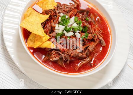 birria de res, stufato di manzo messicano in salsa di peperoncino rosso caldo con cipolla cruda e coriandolo tritato, taco chips in ciotola bianca, primo piano Foto Stock