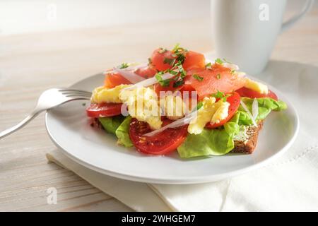 Pane integrale con pomodori, uova strapazzate e salmone affumicato, gustosi sandwich per colazione o pranzo su un luminoso tavolo di legno Foto Stock