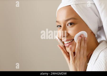 La bella donna con l'abito e l'asciugamano sulla testa pulisce la pelle del suo viso con tamponi di cotone. Pulizia e cura sana del corpo dopo sh Foto Stock