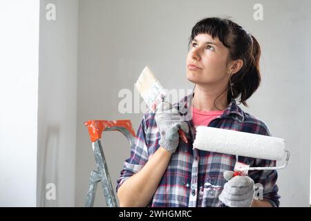 Donna perplessa con rullo per pittura e pittura bianca per pareti in ritratti di primo piano. Lavori di costruzione e riparazioni estetiche in ho Foto Stock