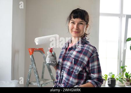 Donna felice con rullo per pittura e pittura bianca per pareti in ritratti da vicino. Lavori di costruzione e riparazioni estetiche in corso Foto Stock