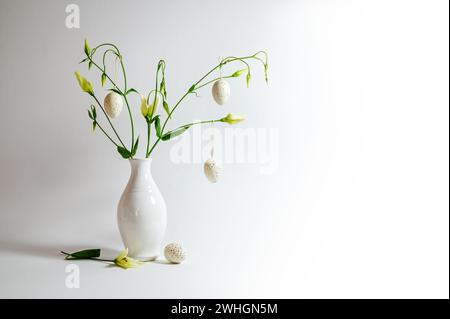 Delicato mazzo di fiori in un vaso bianco decorato con uova di Pasqua appese su sfondo grigio chiaro, spazio copia, messa a fuoco selezionata Foto Stock