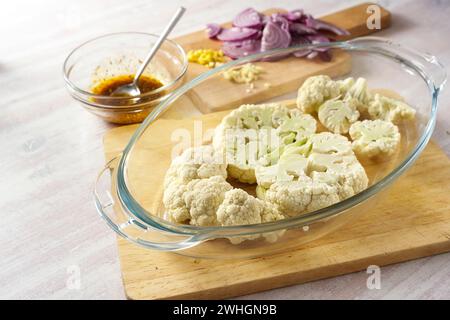 Fette di cavolfiore crude in una casseruola di vetro, cipolla rossa tritata e una miscela di spezie in olio d'oliva, preparazione per la cottura di veget al forno Foto Stock