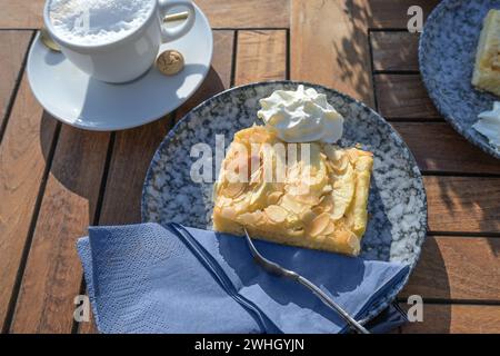 Torta di mele fresca ricoperta di mandorle servita con panna montata su un piatto blu e una tazza di caffè su un tavolo da giardino in legno Foto Stock