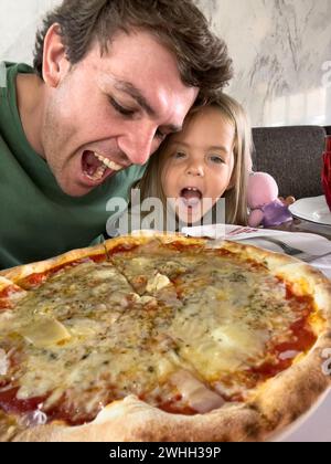 Papà con una bambina che cerca di mordere una pizza grande su un piatto sul tavolo Foto Stock