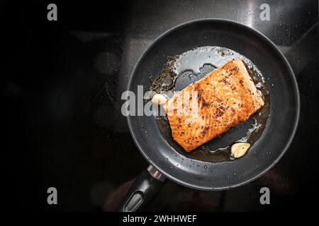 Filetto di salmone fritto con olio d'oliva, erbe aromatiche e aglio in una padella nera sul piano cottura, copia spazio, vista dall'alto Foto Stock