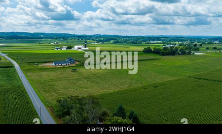 Vista dall'alto Di Una strada curva che attraversa vari campi agricoli con Una casa indipendente e costruzioni adiacenti, sullo sfondo di alberi distanti e cielo pieno di nuvole. Foto Stock