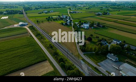Veduta aerea di Una strada curva che corre parallela ai binari ferroviari, fiancheggiata da fattorie e campi, in Un paesaggio agricolo verdeggiante nelle prime ore della sera. Foto Stock