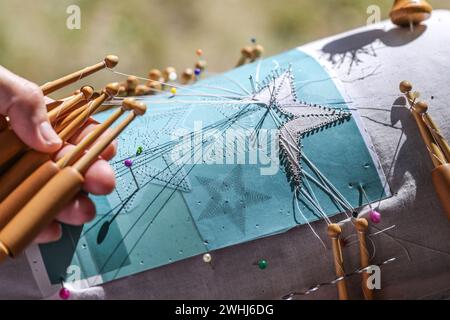 Le mani di una donna che crea una forma a stella di pizzo a bobina con fili su bobine di legno e spille nel motivo sul cuscino in pizzo, deta Foto Stock