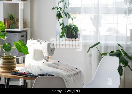 Macchina da cucire su un tavolo rotondo nell'interno bianco della casa vicino alla finestra con una tenda trasparente e un impianto domestico Foto Stock