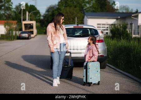 Adorabile bambina si siede sulla valigia del suo bambino e aspetta il viaggio con sua mamma. Due belle ragazze preparano Foto Stock