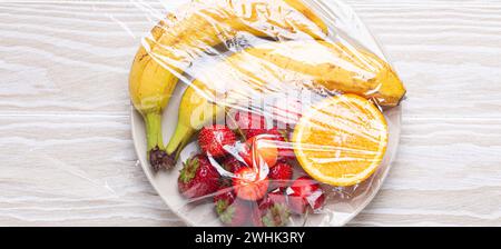 Vari tipi di frutta e frutti di bosco su un piatto bianco avvolto in pellicola trasparente di plastica, vista dall'alto su un tavolo rustico in legno bianco Foto Stock