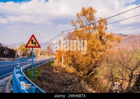 Cartello di avvertimento accanto alla barriera di sicurezza sulla strada di montagna curvata con cielo nuvoloso sullo sfondo in Corea del Sud Foto Stock