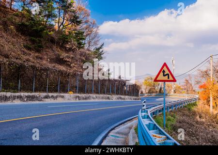 Cartello di avvertimento accanto alla barriera di sicurezza sulla strada di montagna curvata con cielo nuvoloso sullo sfondo in Corea del Sud Foto Stock