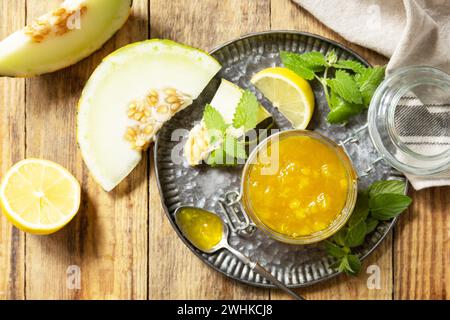 Confettura dolce di melone e agrumi o gelatina in un piccolo vaso di vetro con fette di melone fresche su un tavolo rustico in legno. Riserva naturale fatta in casa. Vista f Foto Stock