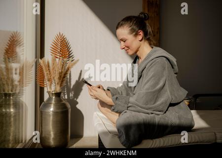 Donna in accappatoio seduta con telefono cellulare in una sala relax durante un pasto benessere Foto Stock