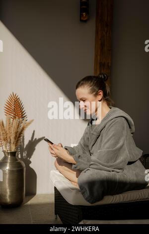 Donna in accappatoio seduta con telefono cellulare in una sala relax durante un pasto benessere Foto Stock