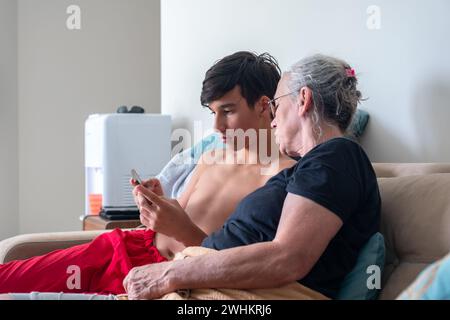 Giovane maschio che insegna sua nonna sui telefoni cellulari moderni e. nuove applicazioni in casa mentre si è seduti in un divano Foto Stock