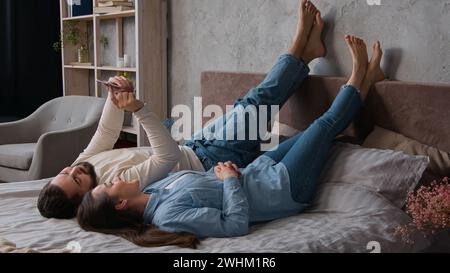 Giovane coppia caucasica spensierata ama rilassarsi sdraiato a letto utilizzando il telefono smartphone gadget. Uomo e donna fanno ordini online giocando su dispositivi mobili Foto Stock