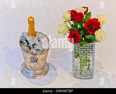 Stile di vita di lusso con bottiglia di vino bianco frizzante in secchio di ghiaccio e mazzo di fiori in un vaso su tovaglia bianca. Foto Stock