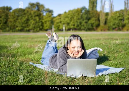 Sorridente ragazza asiatica sdraiata nel parco sull'erba, guardando film o video sul computer portatile, guardando lo schermo con interesse Foto Stock