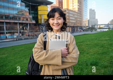 Felice ragazza asiatica si trova sulla strada, studenti universitari cammina con tablet digitale in mani e sorrisi, si trova nel centro della città Foto Stock