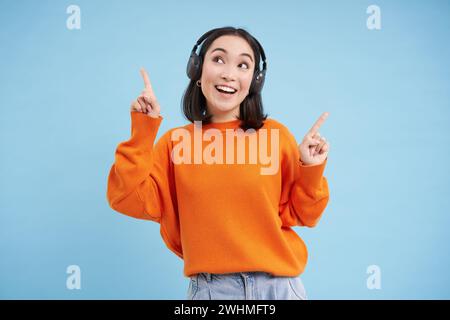 Felice donna cinese in cuffie, ascolta la musica, ama la canzone preferita nella sua playlist, si trova su sfondo blu Foto Stock