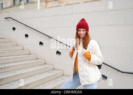 Giovane ragazza felice, turista rossa con cappello rosso con zaino, passeggiate in città, esplorazione della città, zaino in spalla per l'europa, viaggio Foto Stock