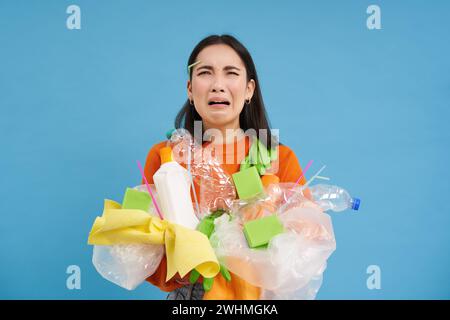 Piangendo femmina asiatica che tiene rifiuti di plastica, spazzatura nelle mani e guardando sconvolto, deve riciclare, sfondo blu Foto Stock