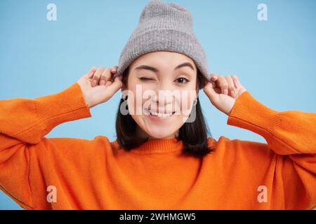 Il ritratto ravvicinato di una modella asiatica indossa il cappello e ti strizza l'occhio, sembrando felice, uscendo, in piedi su un backgroun blu Foto Stock