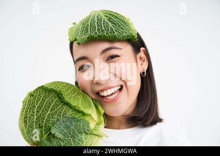 Primo piano ritratto di donna asiatica sorridente sana, che mostra cavolo, letture verdi, con foglia sulla testa, isolato su backgroun bianco Foto Stock