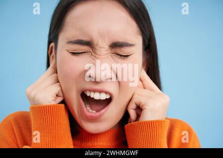 Primo piano ritratto di donna urlante, chiude le orecchie con le dita e grida, si alza su sfondo blu Foto Stock