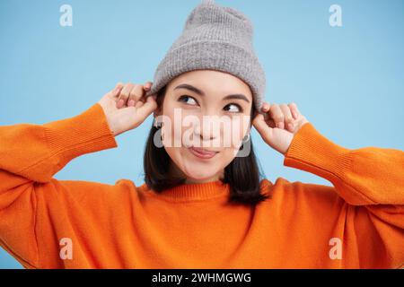 Primo piano ritratto di felice, elegante ragazza coreana mette su caldo cappello, sorride e sembra gioioso, si trova contro sfondo blu studio Foto Stock