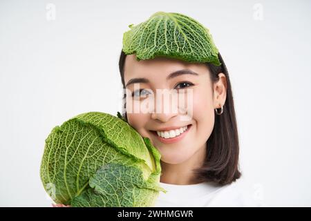 Primo piano ritratto di carina donna asiatica con foglia di lattuga sulla testa, sorridendo e mostrando verde cavolo, sfondo bianco Foto Stock